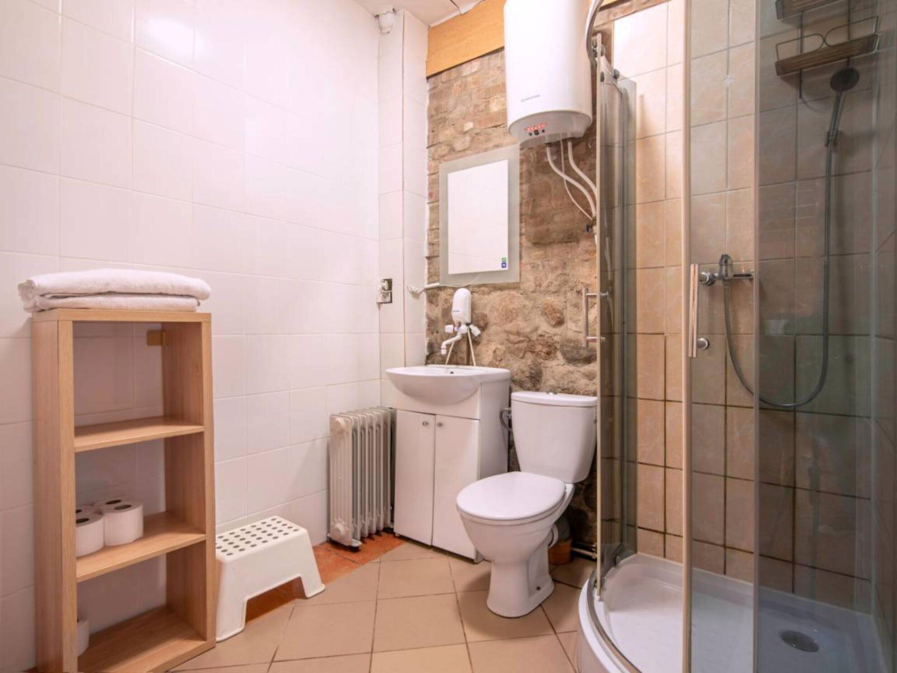 A bathroom with a toilet, sink, shower, and a wooden shelf with towels.