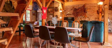 A rustic interior with exposed wooden beams, a dining area with chairs and a table, and a stone wall with a mounted tiger.