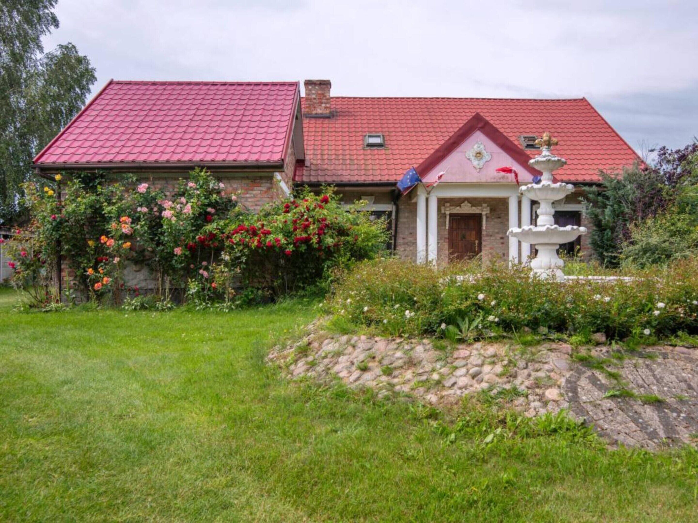 A house with a red roof, a garden with various plants and flowers, and a white fountain.