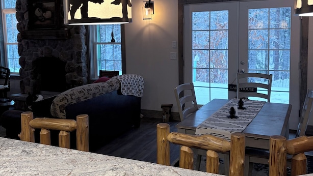 A cozy living room with a stone fireplace, a dining table with chairs, and a large window with a view of trees. - Adorable New 2-bedroom lake house in West Newfields, with Lake access. (West Newfields)