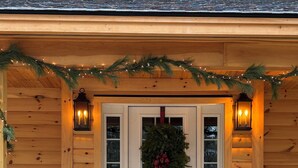 A wooden cabin with a front door decorated with a wreath and lights, a dog sitting on the porch, and a stack of firewood to the left. - Adorable New 2-bedroom lake house in West Newfields, with Lake access. (West Newfields)