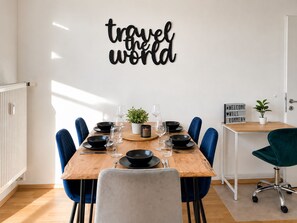 Un coin repas avec une table en bois dressée pour six personnes, entourée de chaises bleues, d’une plante en pot et d’un panneau mural sur lequel on peut lire : « Explorez le monde ».