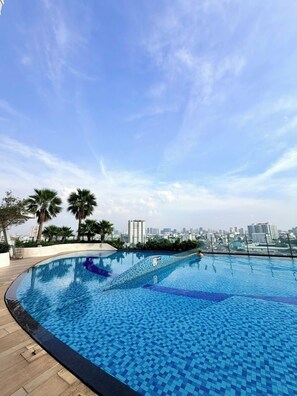 Piscina no terraço com vista para a cidade, rodeada por palmeiras e céu claro.