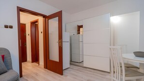 Ett kompakt vardagsrum med en trädörr som leder till en hall, ett vitt matbord med en skål på och en grå soffa med en röd kudde.