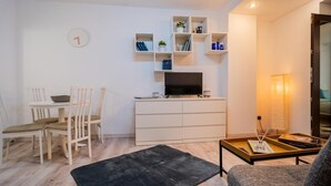 Ett modernt vardagsrum med en matplats, vita möbler, en platt-TV och en liten bokhylla.