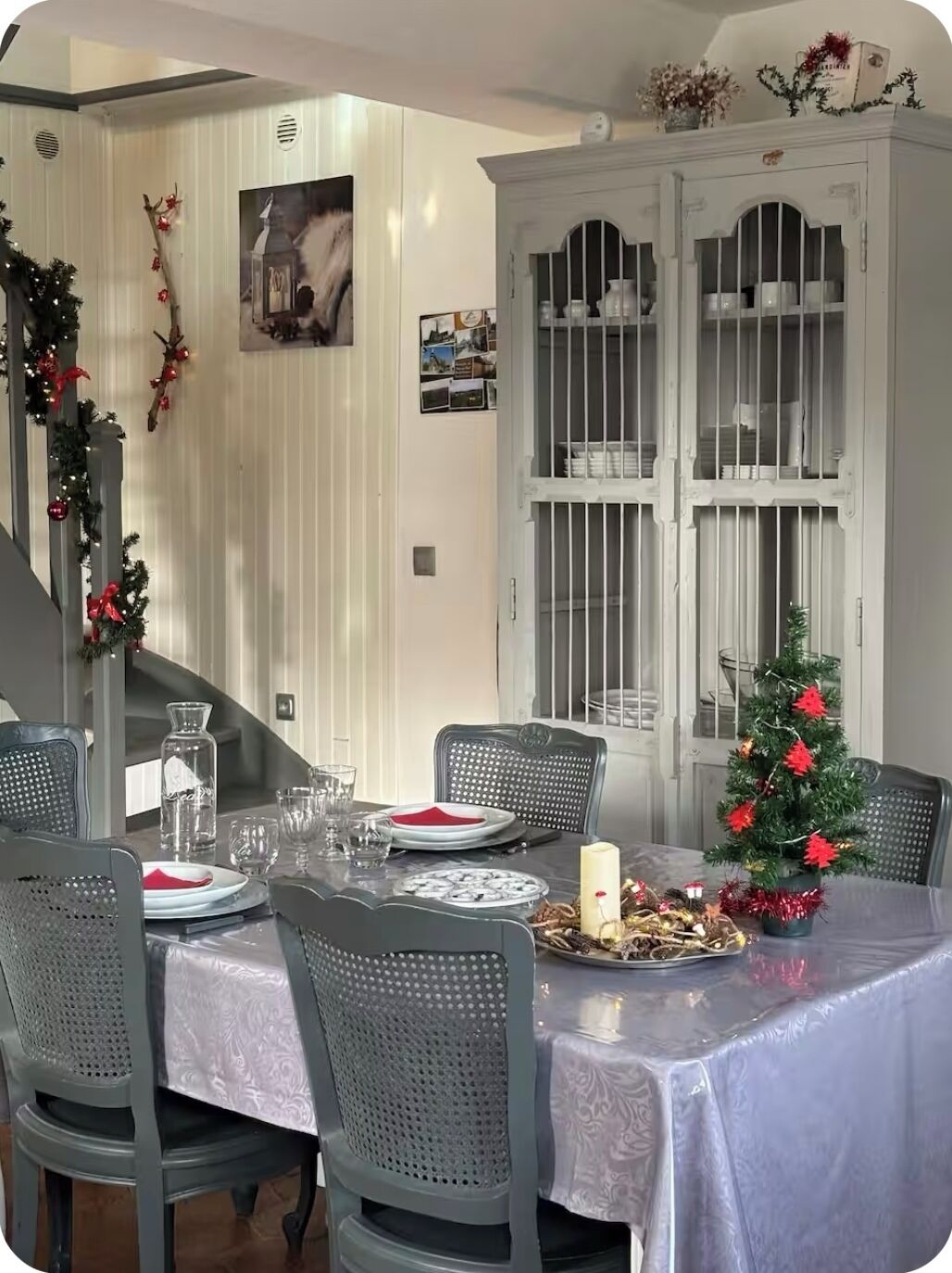 Comedor con una mesa preparada para cuatro personas, rodeada de sillas grises. Un pequeño árbol de Navidad y adornos están sobre la mesa.