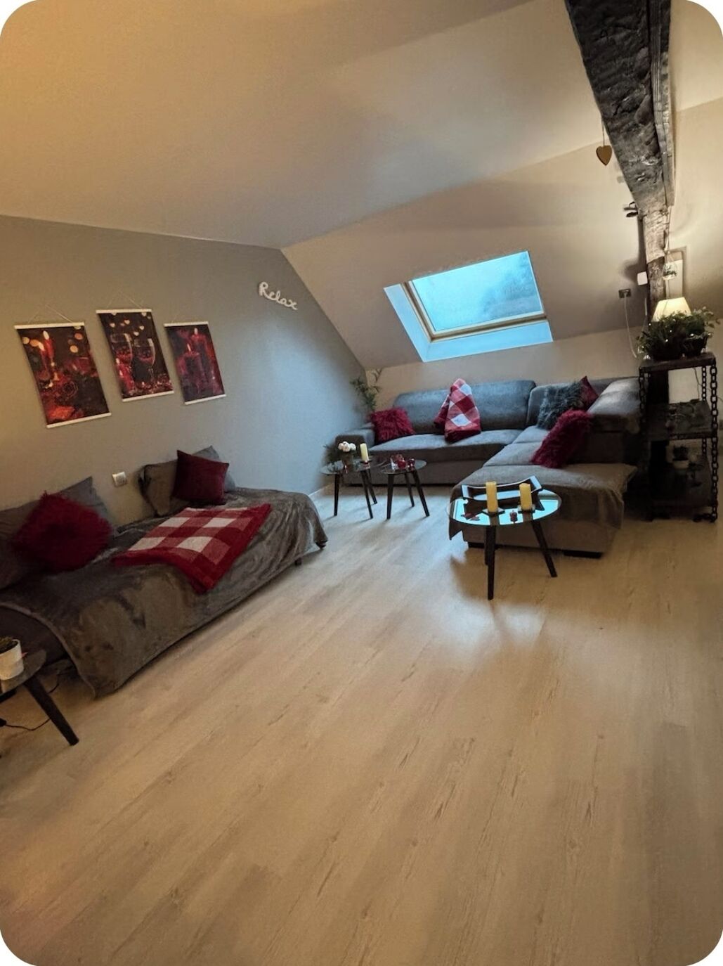 A loft-style living room with a sloped ceiling, a skylight, a grey sofa, a small round table with candles, and three framed pictures on the wall.