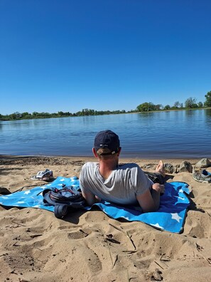 A person sitting on a beach towel by a lake with a clear sky. - ***** Design vacation home Cameroon on the Elbe "Casa Kamerunschaf" (Damnatz)
