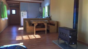 Una habitación pequeña con una mesa y banco de madera, una kitchenette con refrigerador y un estufa a leña.