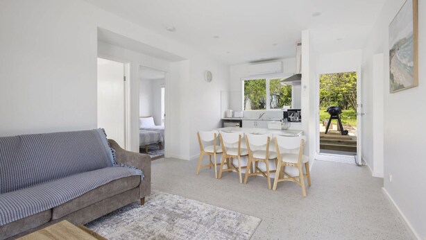 A modern living room with a grey sofa and a wooden coffee table, a dining area with white chairs, and a kitchen with white cabinets and a window. - Peaceful 2-bed Apartment in Coastal Mccrae (Melbourne)