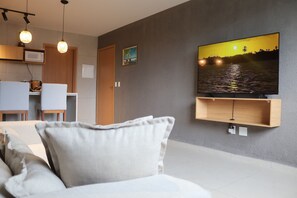 A modern living room with a flat-screen TV mounted on a grey wall, a wooden cabinet below it, and a dining area with a microwave and chairs. - MHV Milagres - Manah B 212 &  B 211  (São Miguel dos Milagres)