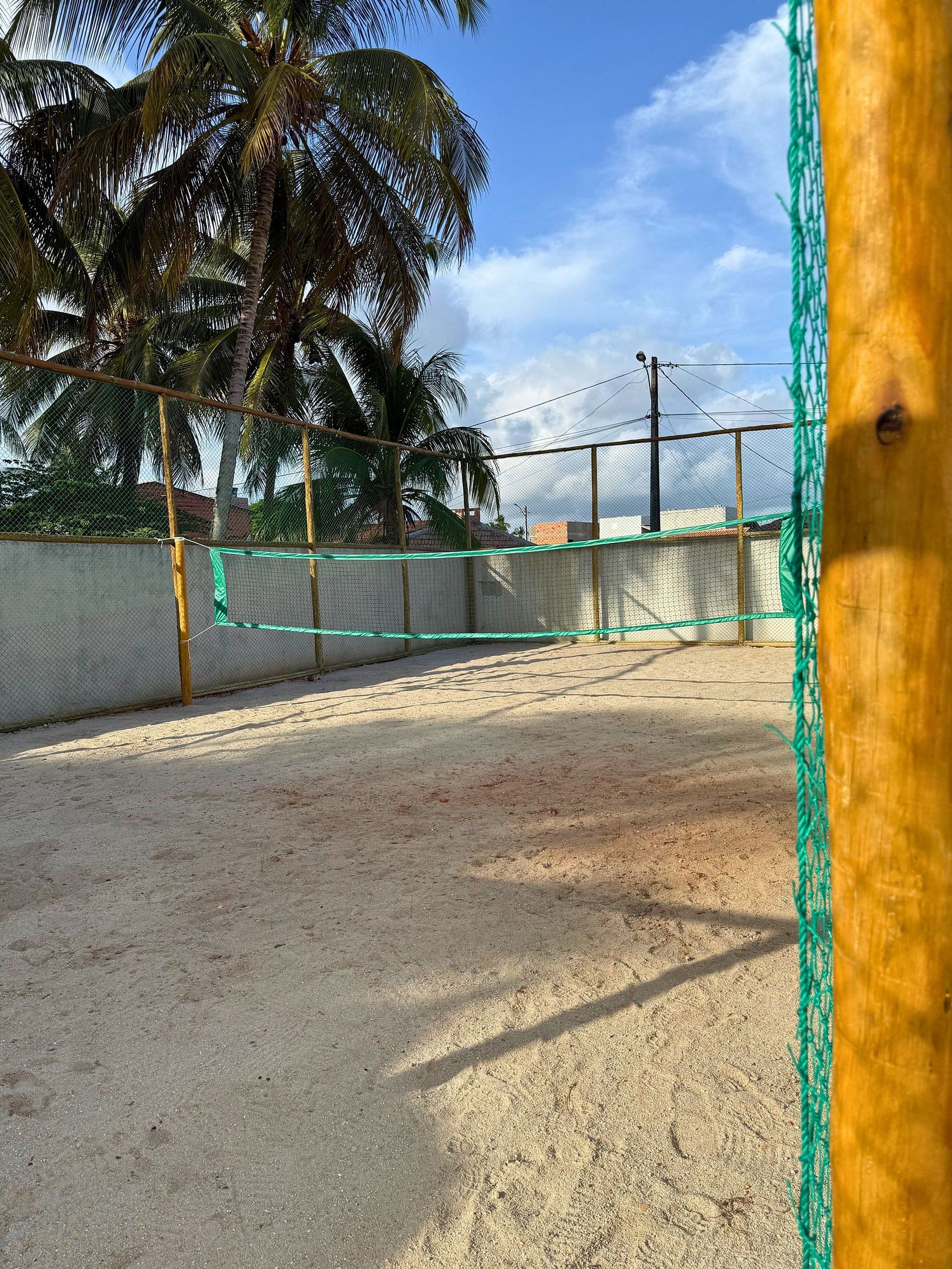 A sandy outdoor area with a volleyball net, palm trees, and a fence.
