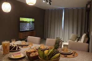A living room with a flat-screen TV, a dining table set with food and drinks, a sofa, and a large window with curtains. - MHV Milagres - Manah B 212 &  B 211  (São Miguel dos Milagres)