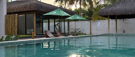 Una piscina con lettini e ombrelloni, circondata da palme e costruzioni con tetti di paglia.
