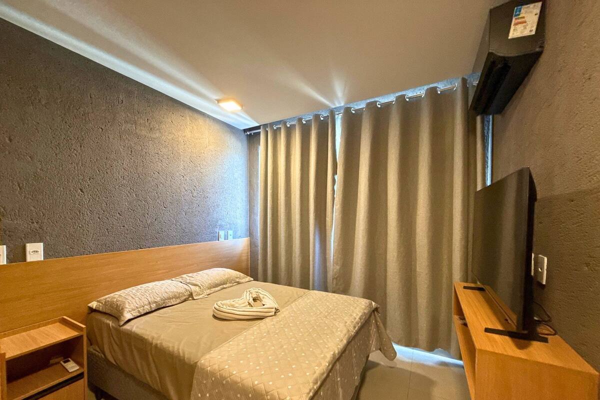 A hotel room with a bed, a TV, a wooden nightstand, and a window with curtains.