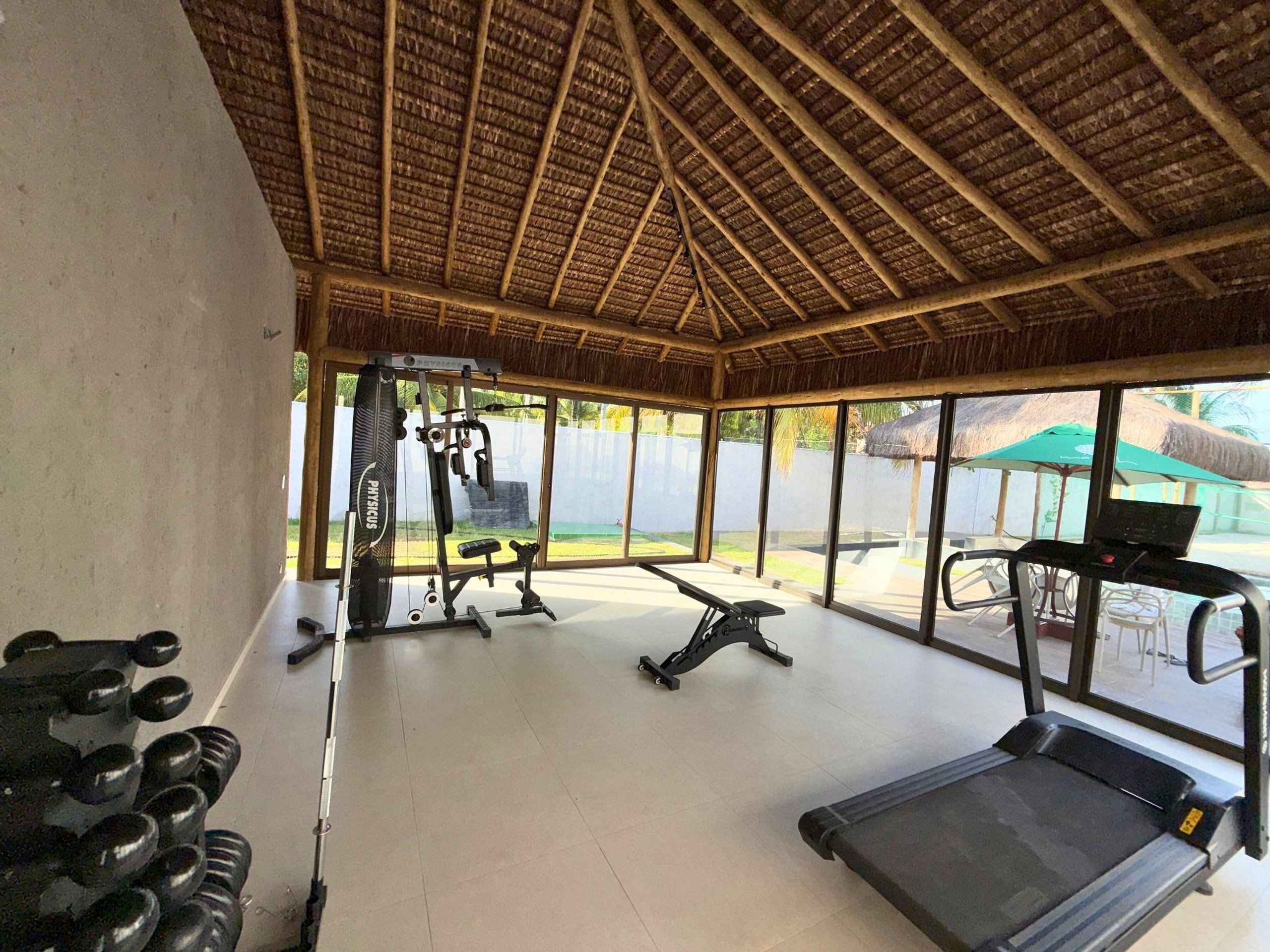 Ein Fitnessraum mit Trainingsgeräten, Laufband und großen Fenstern mit Blick auf einen Garten.