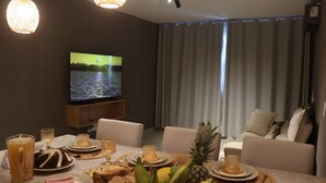 Un soggiorno con televisione a schermo piatto, un tavolo da pranzo apparecchiato con cibo e bevande, un divano e un'ampia finestra con tende.