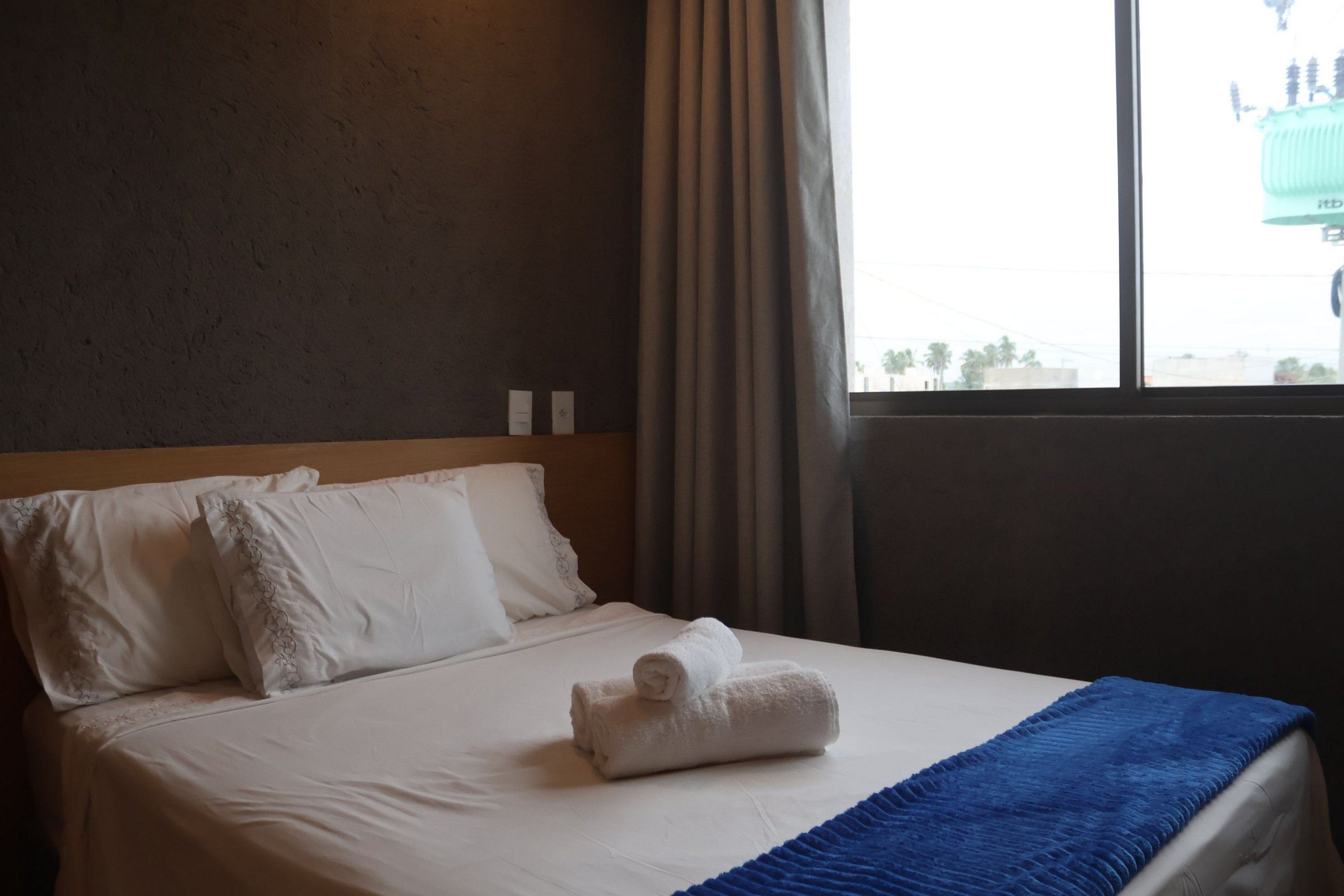 A hotel room with a bed, white linens, a blue blanket, and a window with curtains.