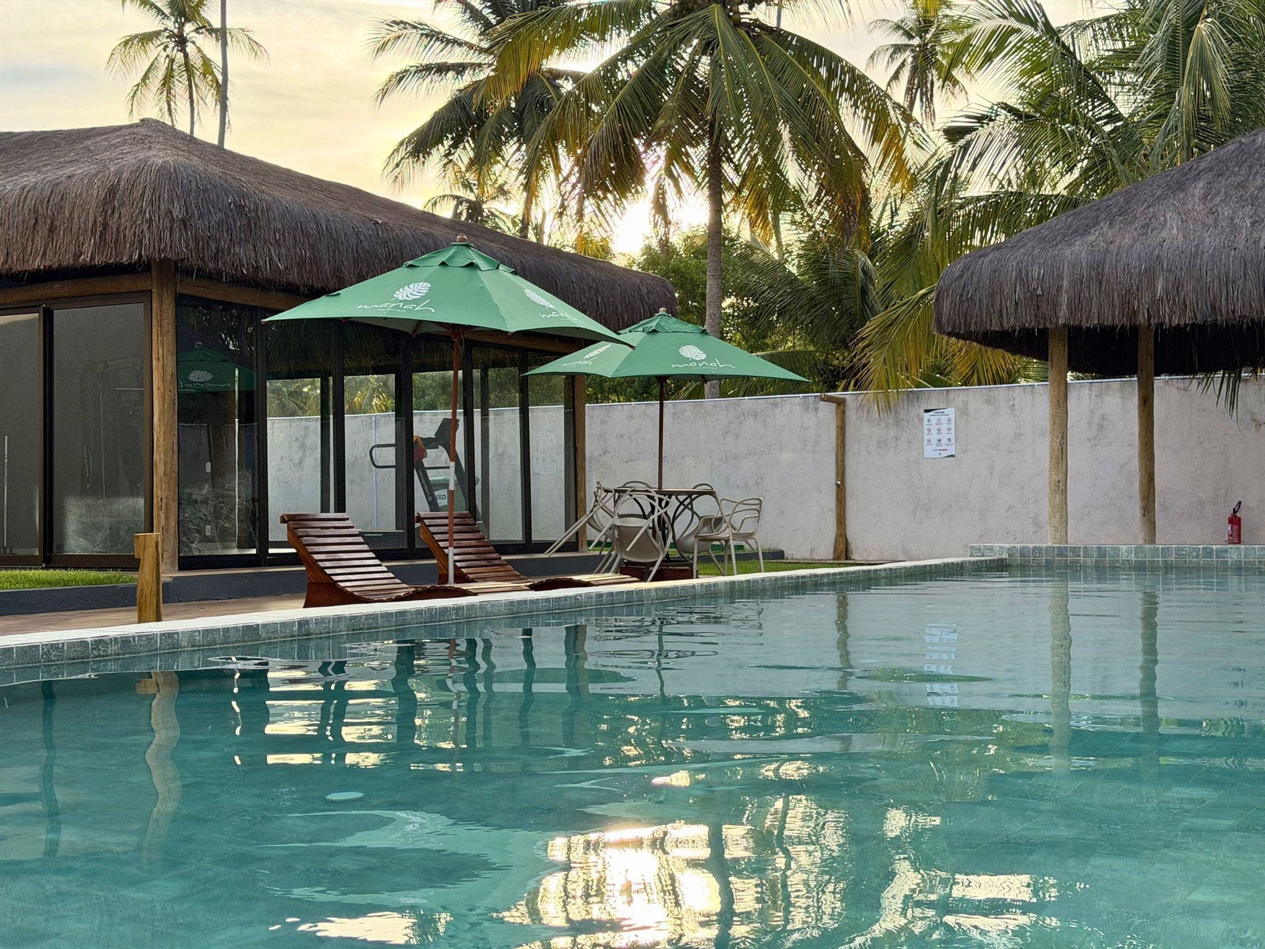 Une piscine entourée de palmiers et dotée d’une structure à toit de chaume, avec des chaises longues et des parasols à proximité.