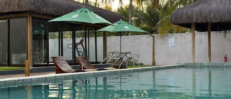 A swimming pool with lounge chairs and umbrellas, surrounded by palm trees and a thatched-roof structure.