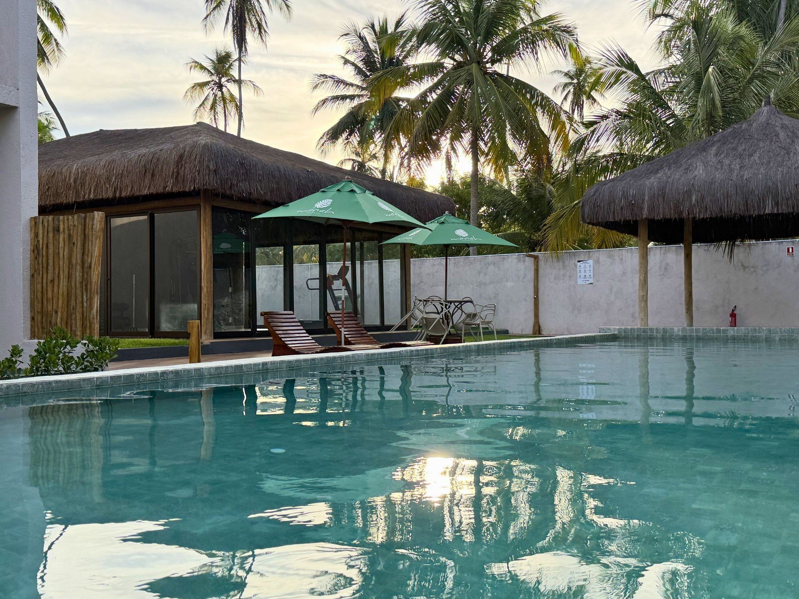 Ein Swimmingpool mit Liegestühlen und Sonnenschirmen, umgeben von Palmen und strohgedeckten Hütten.