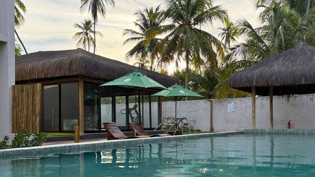 Una piscina con lettini e ombrelloni, circondata da palme e costruzioni con tetti di paglia.