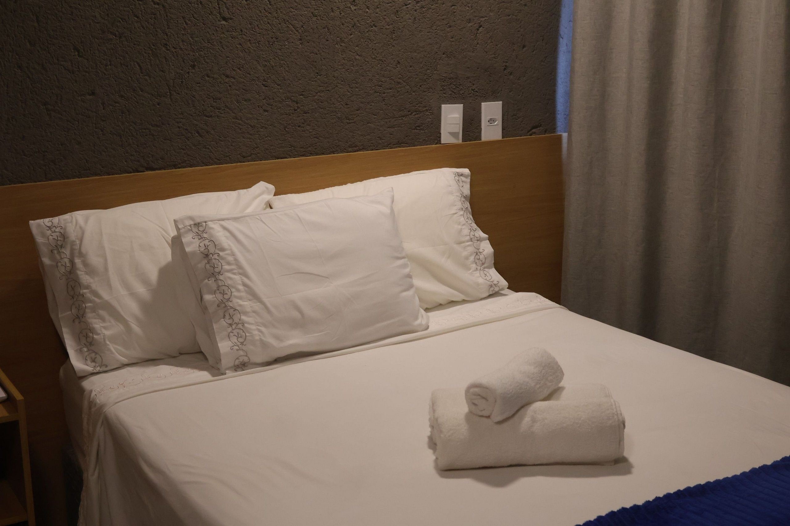 Ein ordentlich bezogenes Bett mit weißen Leinen und einem gefalteten Handtuch, ein Holz-Kopfteil und eine schlichte Wand mit einer Steckdose.