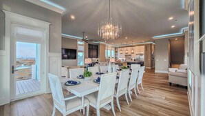 A spacious dining area with a long table and white chairs, a chandelier, and a view of the kitchen and living area. - Tide Chateau – Epic Sunsets + Golf Cart Access! (Surf City)