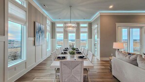 A spacious dining area with a long table, white chairs, and a chandelier. Large windows offer views of the landscape. - Tide Chateau – Epic Sunsets + Golf Cart Access! (Surf City)