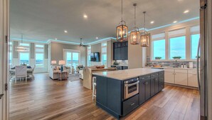 A spacious kitchen with a large island, modern pendant lighting, and a view of the ocean. - Tide Chateau – Epic Sunsets + Golf Cart Access! (Surf City)