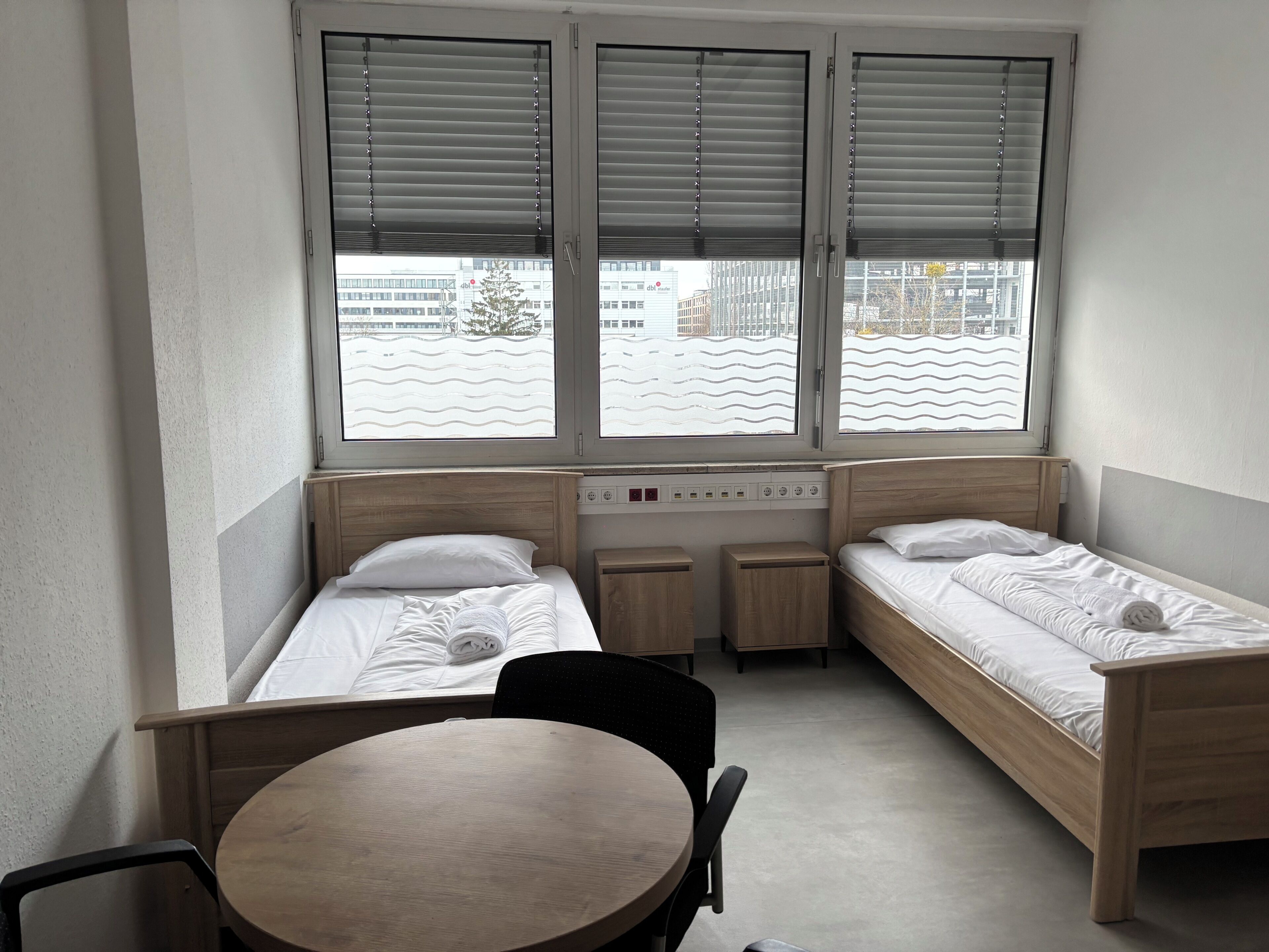 A small, modern room with two single beds, a round table, and a chair.
