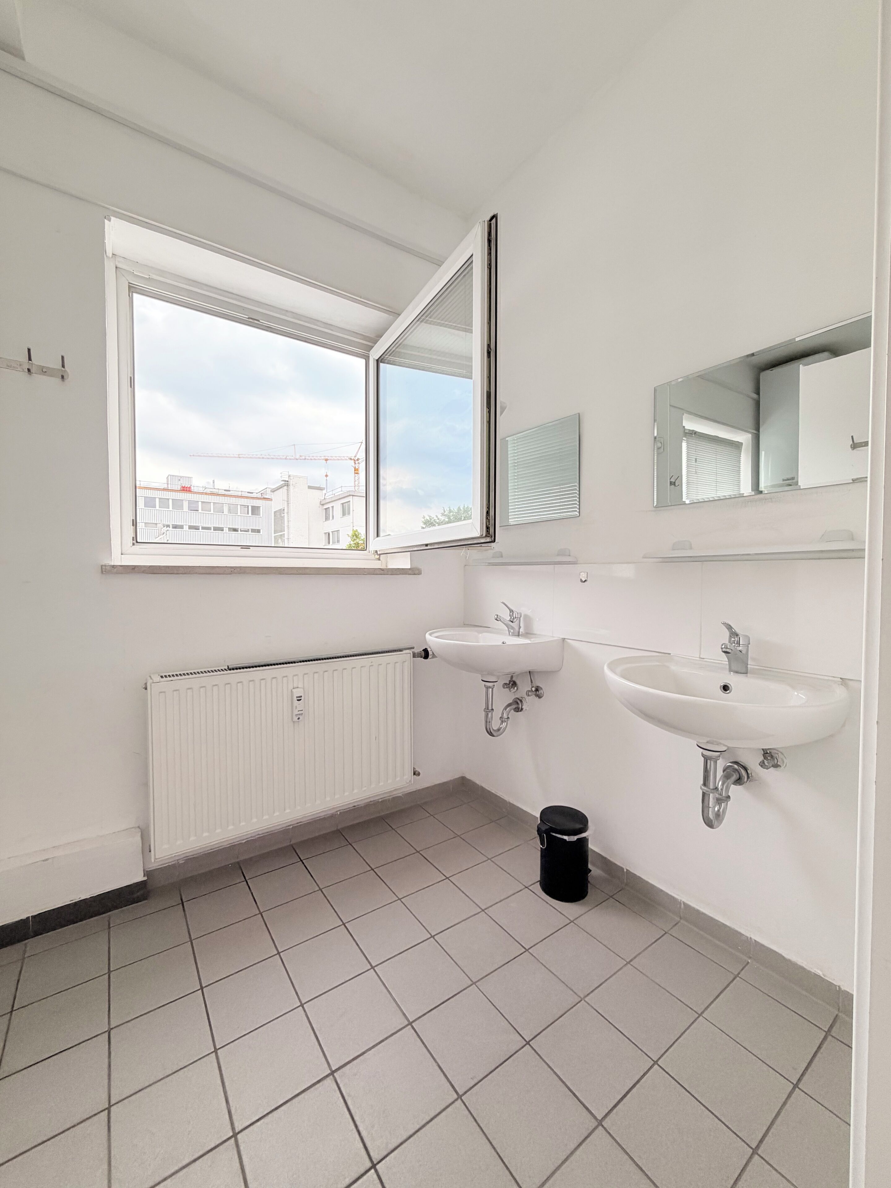 A bathroom with two sinks, a window, a radiator, and a black trash bin.