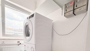 A laundry room with a front-loading washer and dryer stacked on top of each other, a window with a view of buildings, and a wall-mounted electrical outlet. - Haus an der Brücke (Munich)