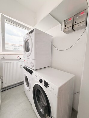 A laundry room with a front-loading washer and dryer stacked on top of each other, a window with a view of buildings, and a wall-mounted electrical outlet. - Haus an der Brücke (Munich)