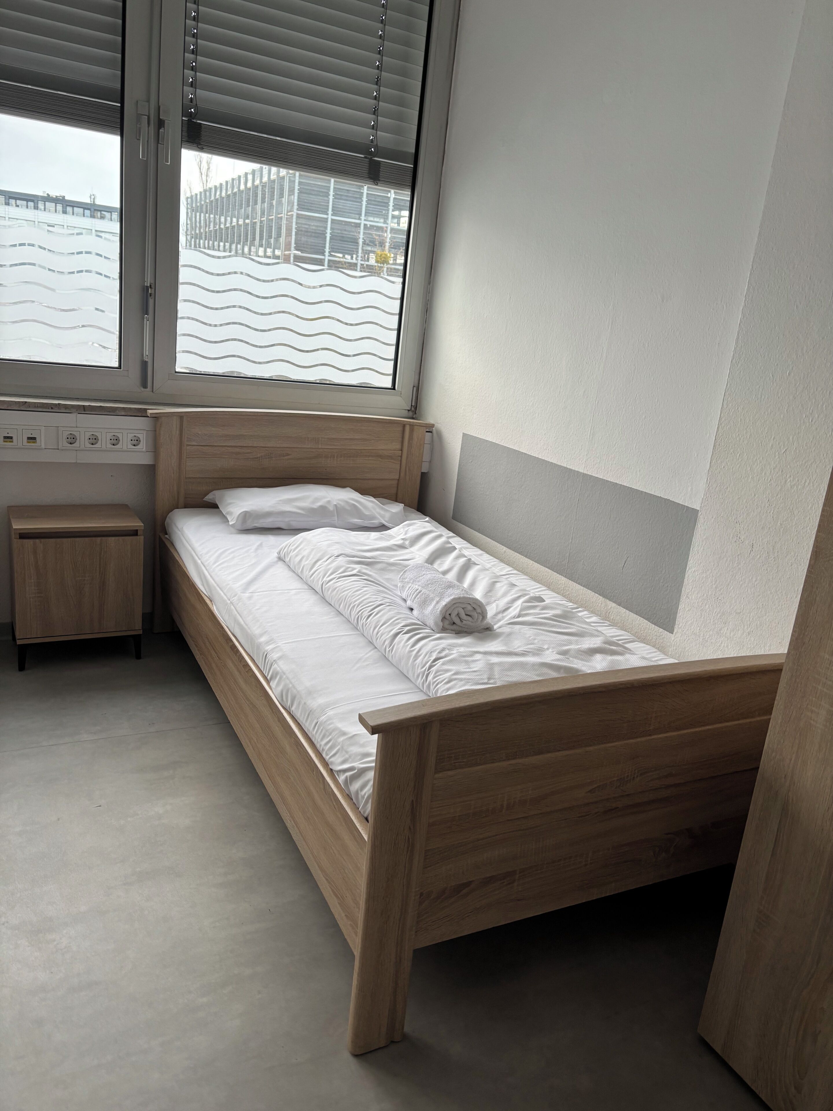 Ein Einzelbett mit Holzrahmen und weißer Bettwäsche, ein kleiner Nachttisch und ein Fenster mit Jalousien.