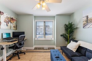 A room with a desk, two monitors, a chair, a sofa, a large window, a ceiling fan, and a plant. - Beautifully Updated Diamond Beach Townhouse (Wildwood)