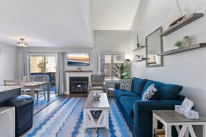 A living room with a sofa, coffee table, and fireplace. There is a dining area with a table and chairs. The room has a view of the outdoors. - Beautifully Updated Diamond Beach Townhouse (Wildwood)