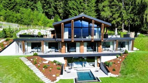Une maison moderne à deux étages, dotée d’une grande façade vitrée, d’une piscine et d’un mur de soutènement en pierre.