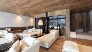 Une chambre d’hôtel moderne avec un plafond en bois, une grande fenêtre offrant une vue sur les montagnes et un coin salon confortable avec des canapés et une table basse.