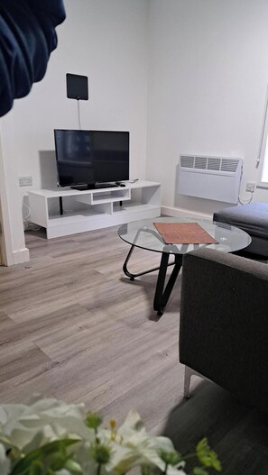 A modern living room with a flat-screen TV on a white entertainment unit, a glass coffee table, and a grey sofa. - Central Sheffield 1-Bed | Walk to Shops & Dining (Sheffield)