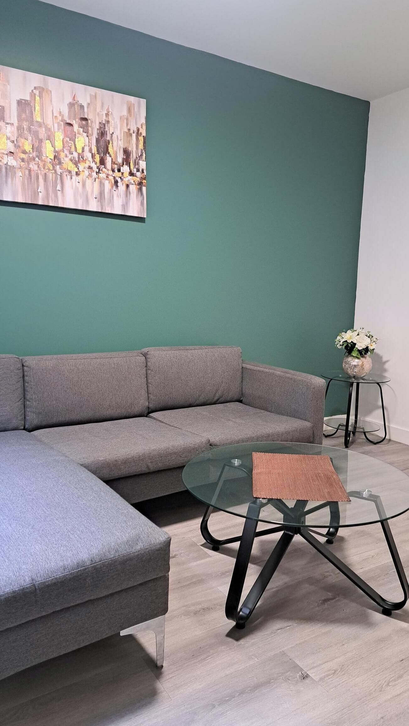 Un salon moderne avec un canapé d’angle gris, une table basse en verre surmontée d’un set de table en fibres tressées, et un tableau représentant un paysage urbain accroché au mur de couleur turquoise.