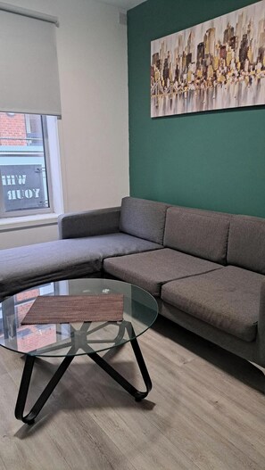 A modern living room with a grey sectional sofa, a glass coffee table, and a cityscape painting on the wall. - Central Sheffield 1-Bed | Walk to Shops & Dining (Sheffield)