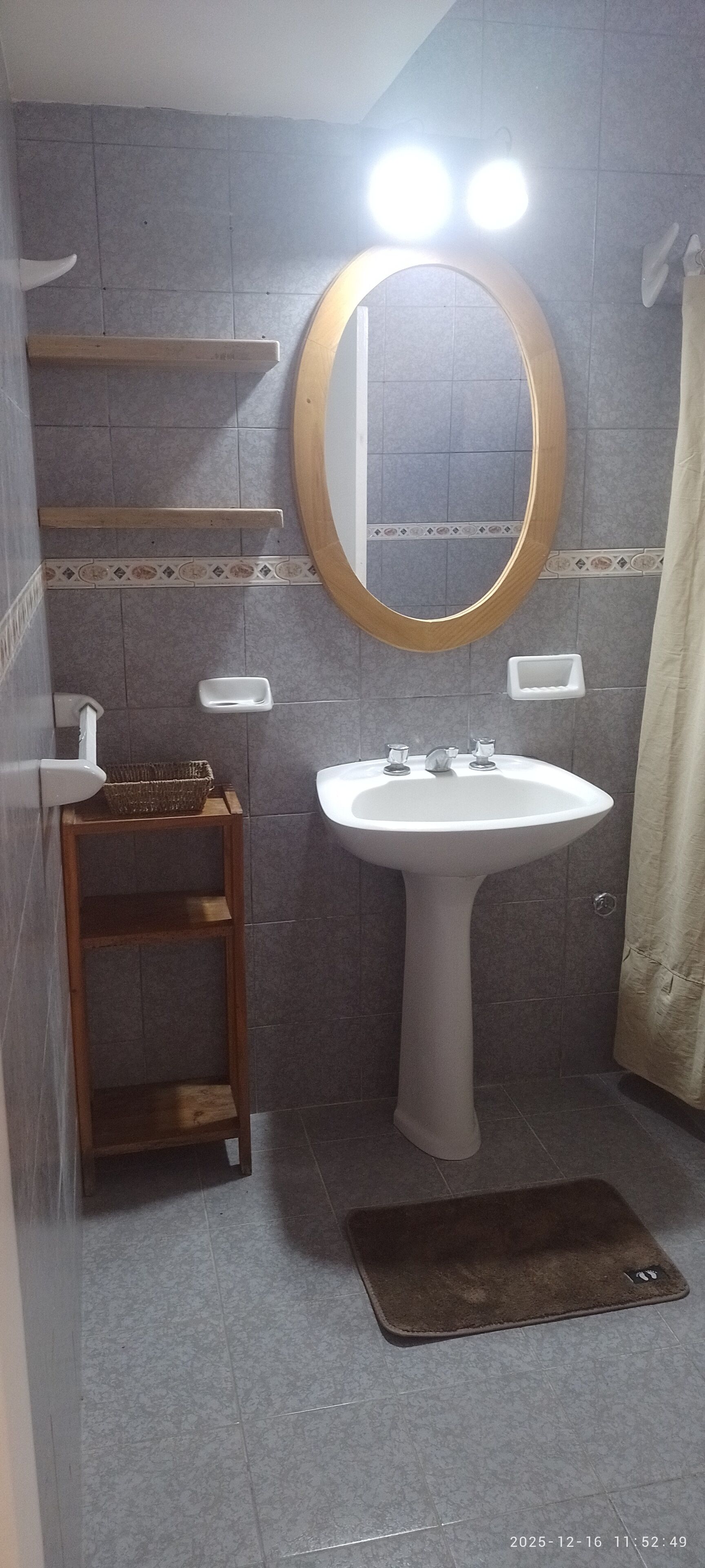Une salle de bain avec un lavabo sur colonne, un miroir avec un cadre en bois, une petite étagère et un rideau de douche.