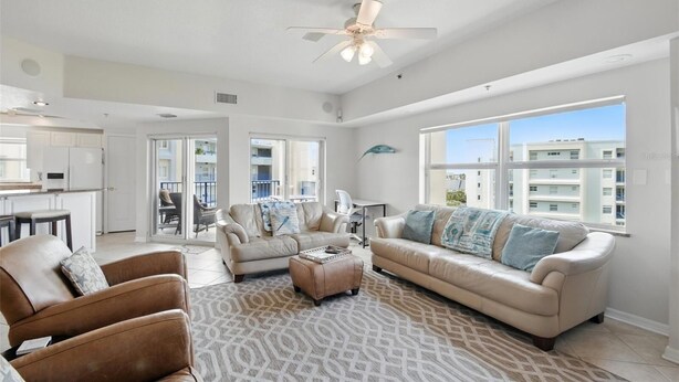 Un salon spacieux avec un canapé beige, deux fauteuils, une table basse et un tapis à motifs. On y trouve un ventilateur de plafond, une grande fenêtre donnant sur des immeubles et une porte menant à un balcon.