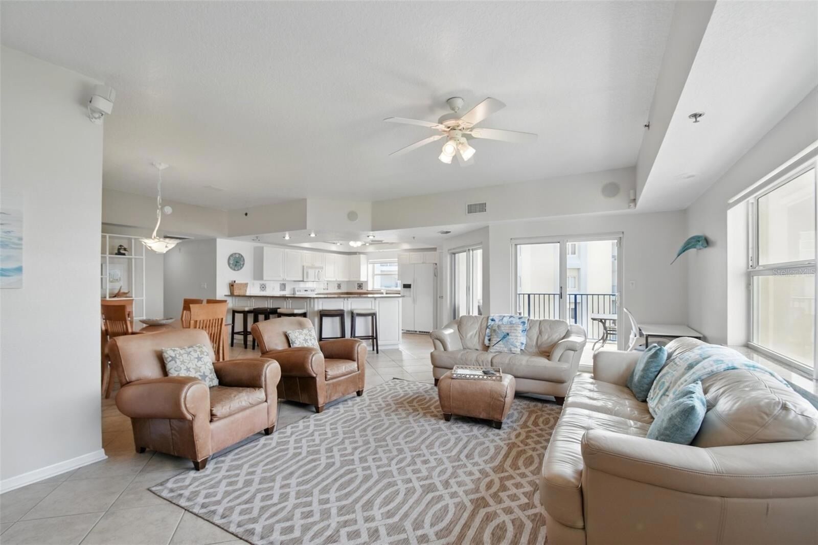 A spacious living room with a large sofa, two armchairs, a coffee table, and a patterned rug. There is a kitchen in the background with white cabinets and a dining area with wooden chairs.