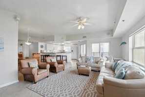A spacious living room with a large sofa, two armchairs, a coffee table, and a patterned rug. There is a kitchen in the background with white cabinets and a dining area with wooden chairs.