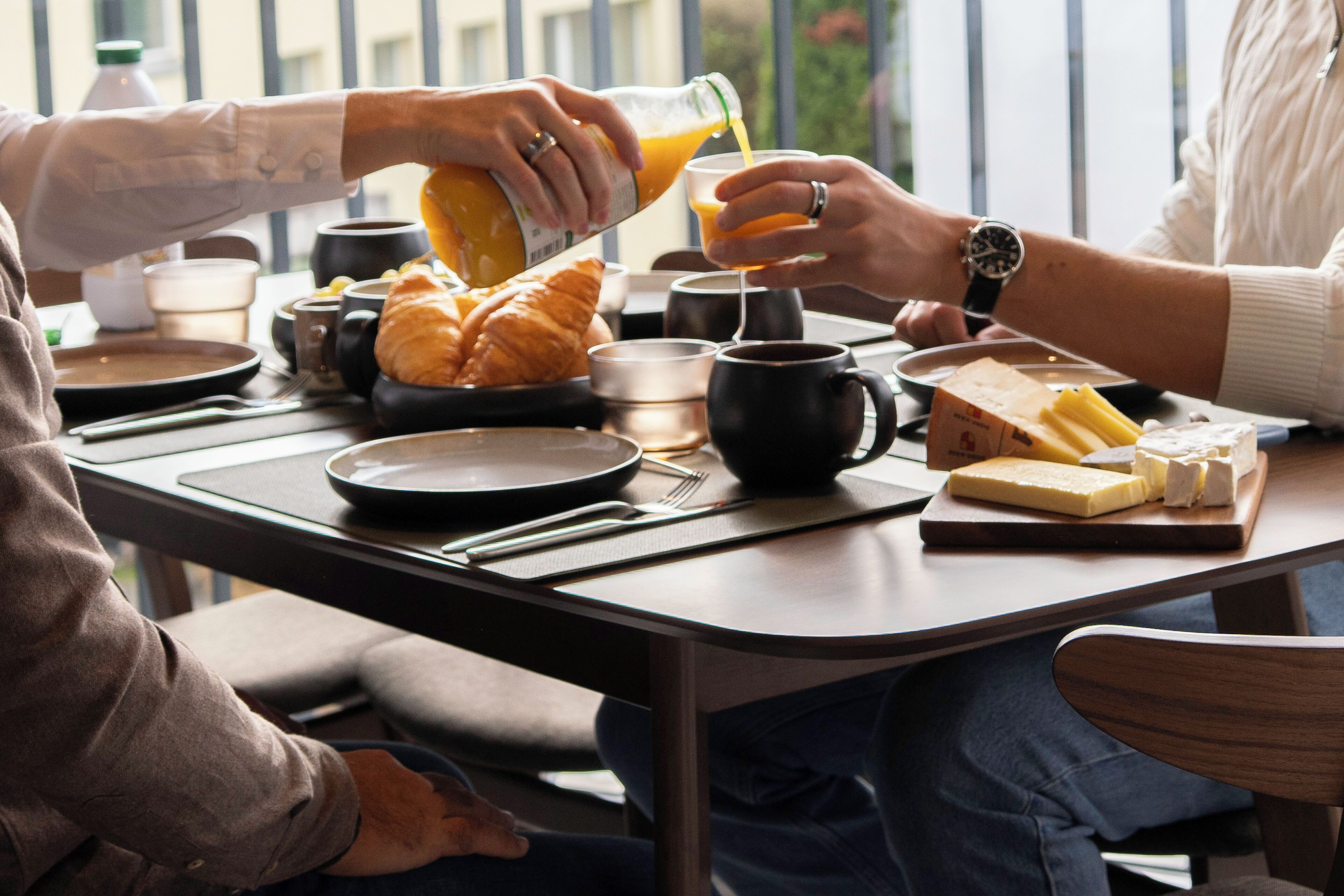 Una mesa de desayuno lista para dos personas, con croissants, queso y una botella de jugo de naranja.
