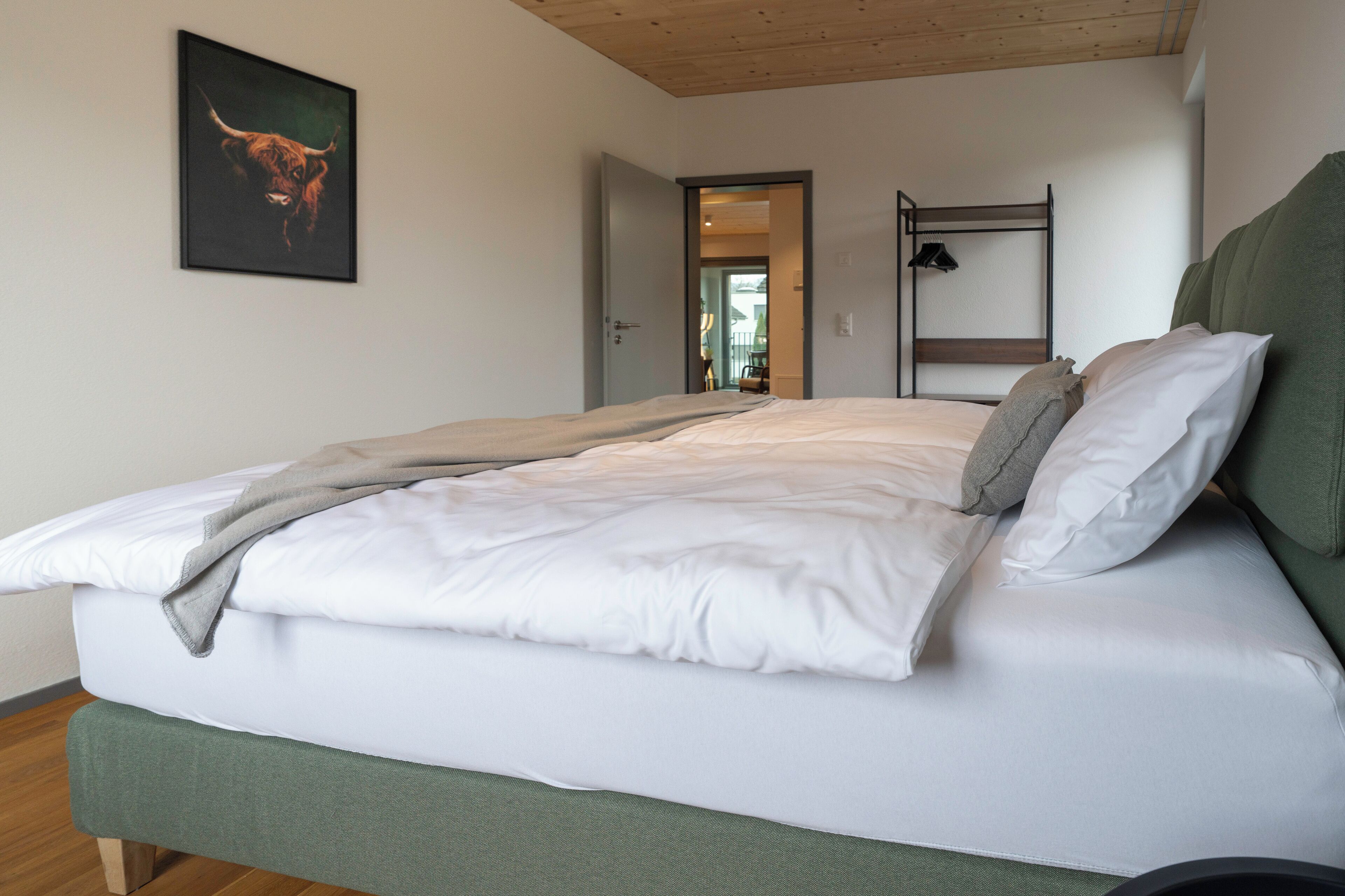 Ein ordentlich bezogenes Bett mit weißen Leinen und einem grünen Kopfteil, ein Holzbett im Hintergrund und ein Bullenbild an der Wand.