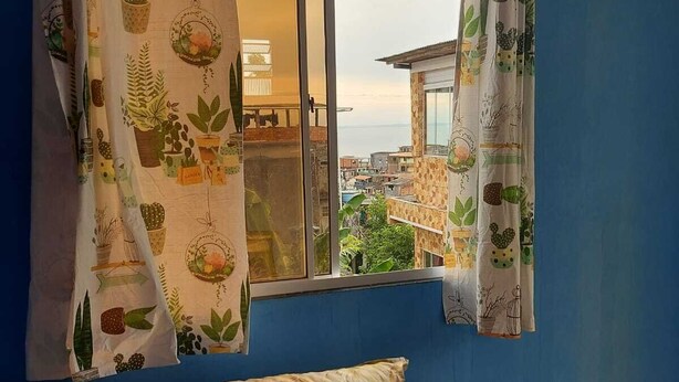 A room with a bed, a window with curtains, and a view of buildings and greenery outside. - House, 2 bedrooms with air conditioning, airy, fully equipped. (Salvador)