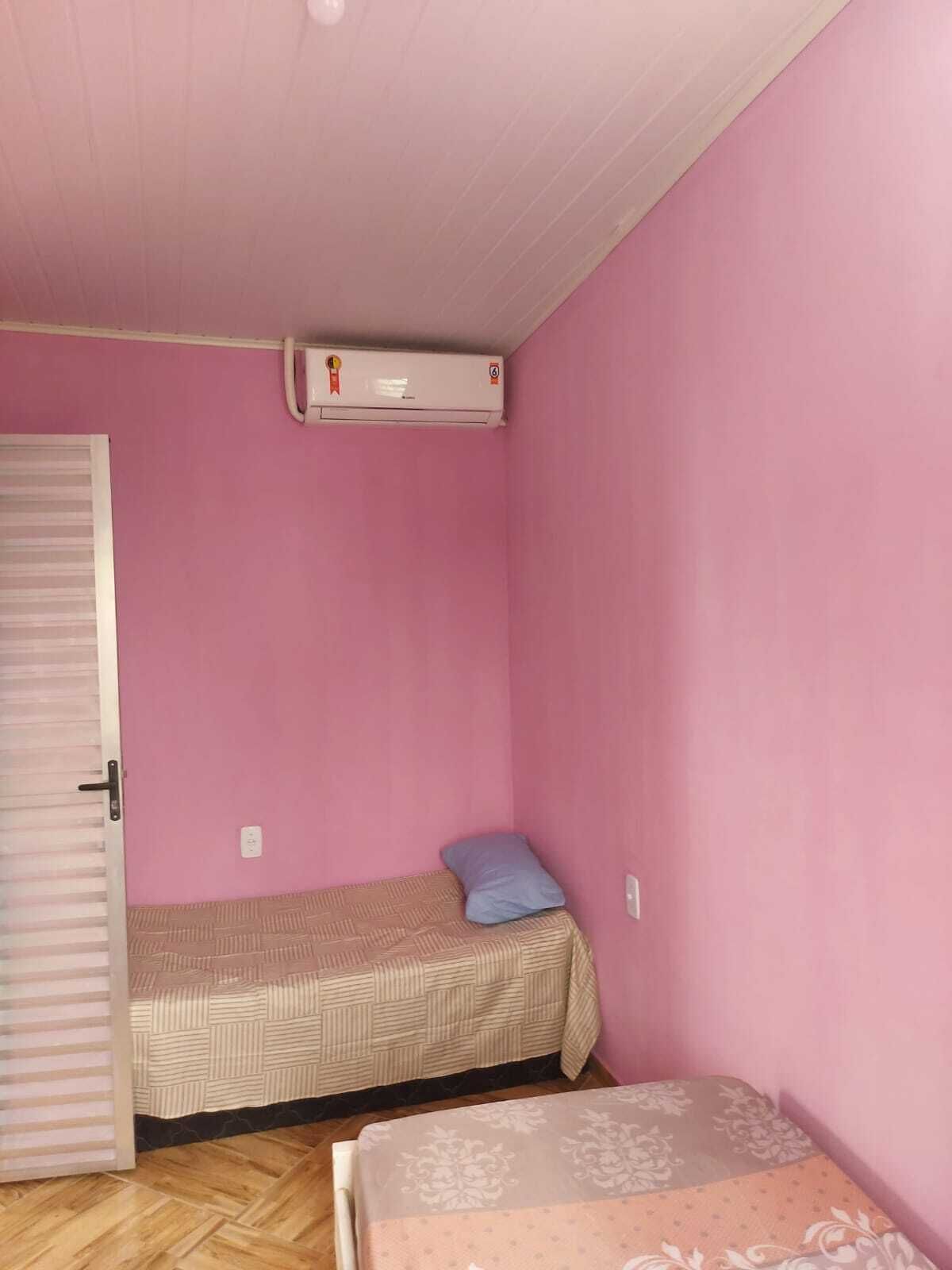 Une petite pièce aux murs roses, avec un lit simple, une fenêtre avec des stores et un climatiseur fixé au mur.